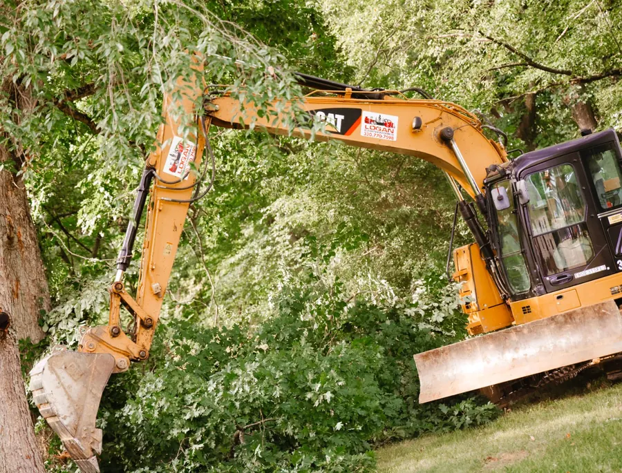 CAT excavator with Logan Earthworks decals reaching its bucket into dense trees